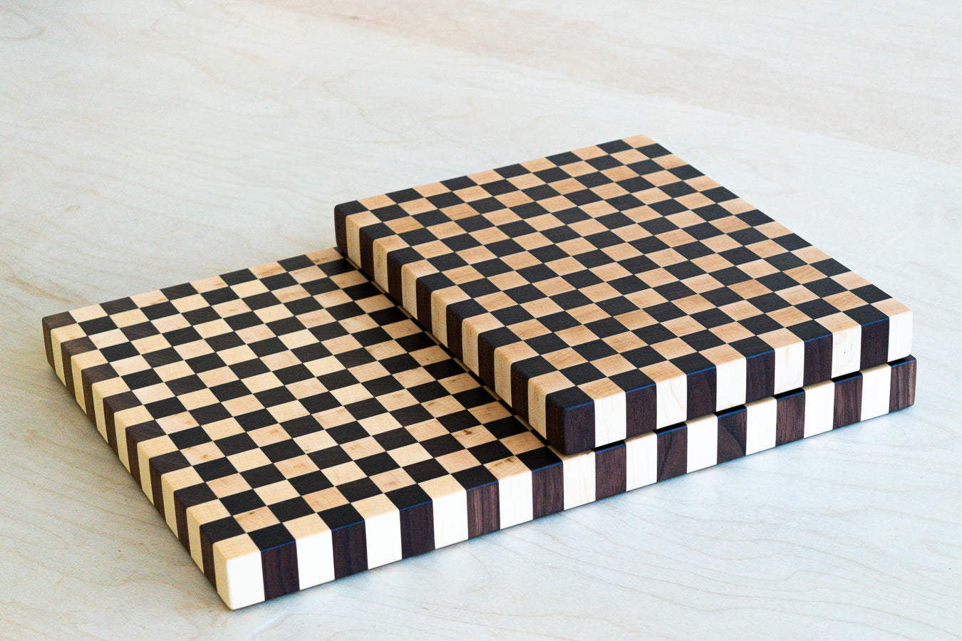 Two cutting boards with a checked patern stacked on top of one another, the top one is smaller. On a light wood background.